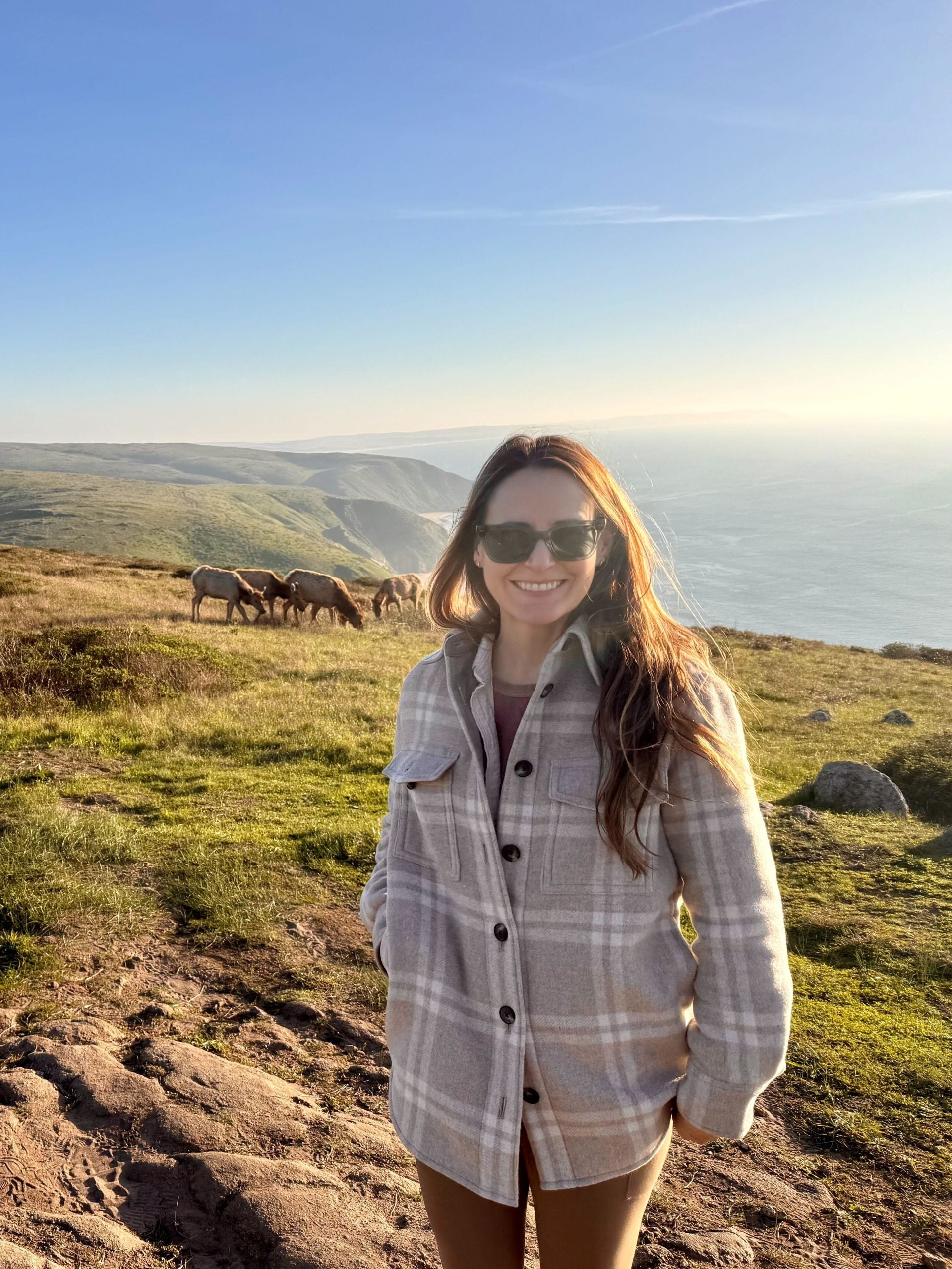 Kristen, smiling, stands atop an oceanside cliff near a herd of Tule elk